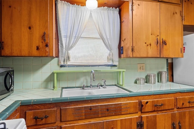 kitchen with tile countertops, brown cabinets, a sink, black microwave, and backsplash