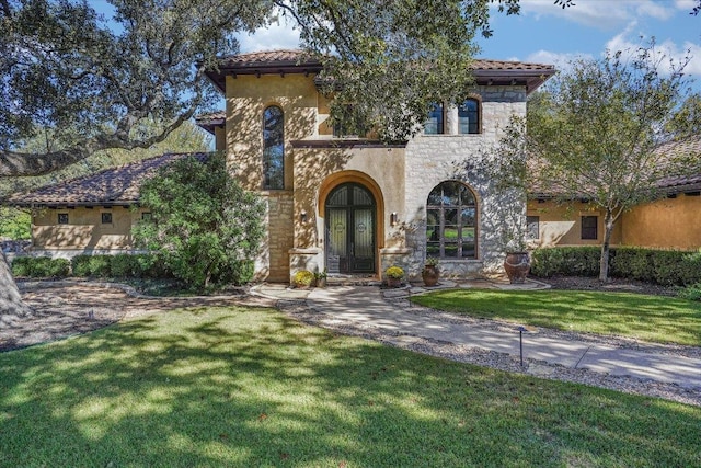 mediterranean / spanish home featuring a tile roof, stone siding, french doors, stucco siding, and a front lawn