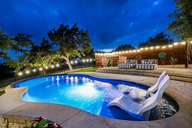 pool at night featuring a patio, a water slide, a fenced backyard, and a fenced in pool