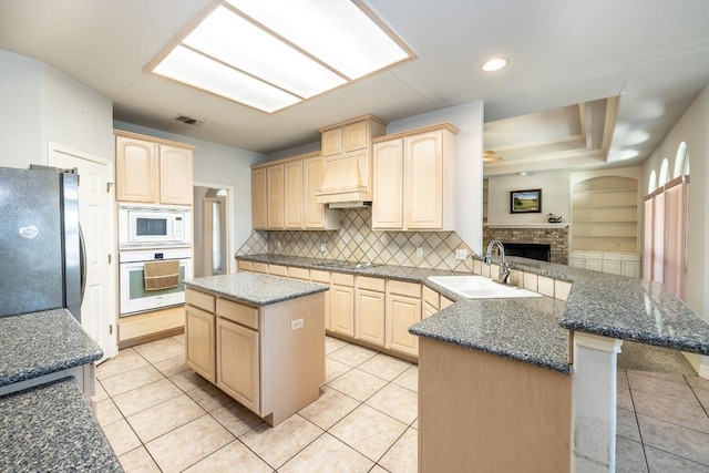 kitchen featuring a center island, light brown cabinetry, a sink, white appliances, and a peninsula