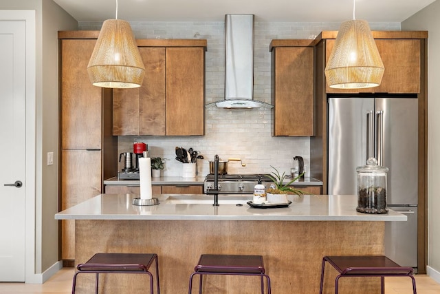 kitchen with light countertops, decorative backsplash, freestanding refrigerator, brown cabinets, and wall chimney exhaust hood