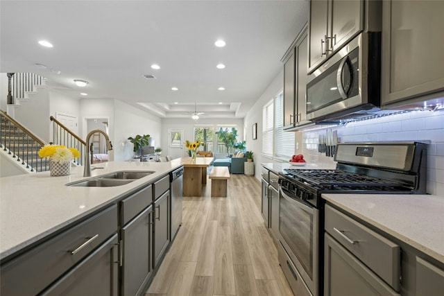 kitchen featuring appliances with stainless steel finishes, light stone counters, a tray ceiling, light wood-style floors, and a sink