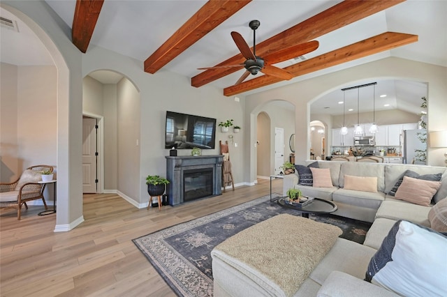 living room featuring baseboards, a ceiling fan, a glass covered fireplace, vaulted ceiling with beams, and light wood-type flooring