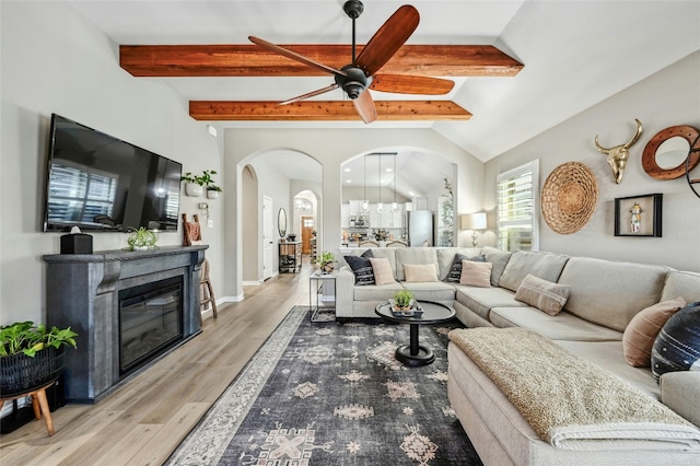 living room with arched walkways, a ceiling fan, a glass covered fireplace, vaulted ceiling with beams, and light wood-type flooring