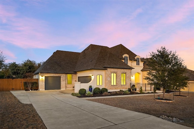 french country home featuring a garage, concrete driveway, roof with shingles, and fence