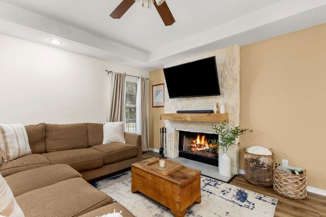 living room featuring baseboards, ceiling fan, wood finished floors, a tray ceiling, and a stone fireplace