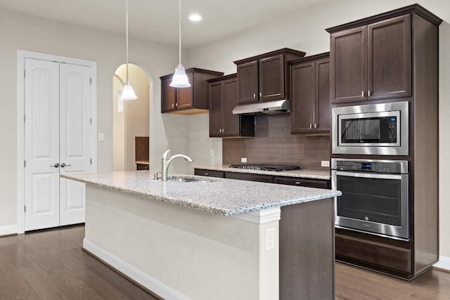 kitchen featuring under cabinet range hood, a sink, appliances with stainless steel finishes, light stone countertops, and decorative light fixtures