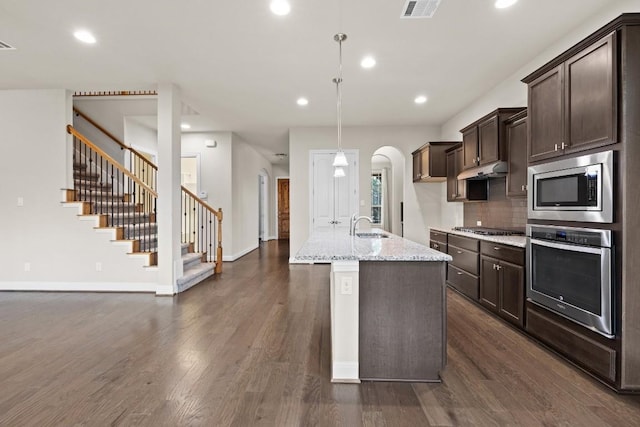 kitchen featuring light stone counters, pendant lighting, dark wood finished floors, appliances with stainless steel finishes, and an island with sink