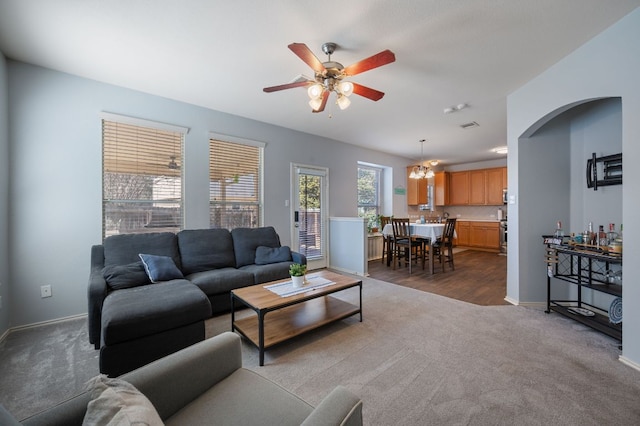 living area featuring arched walkways, dark colored carpet, ceiling fan with notable chandelier, and baseboards