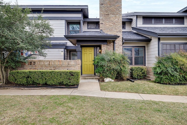 entrance to property with a shingled roof, a chimney, a lawn, and brick siding