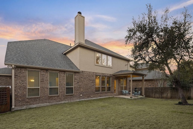 back of house at dusk featuring a shingled roof, fence, a yard, a chimney, and a patio area