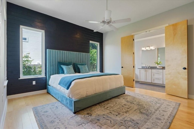 bedroom featuring ceiling fan, wood walls, baseboards, light wood-type flooring, and ensuite bath