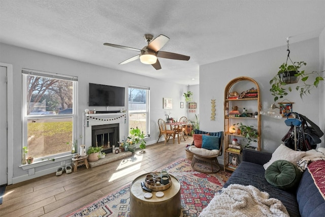 living area with a fireplace with raised hearth, ceiling fan, a textured ceiling, wood finished floors, and baseboards