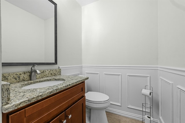 half bath with toilet, a decorative wall, a wainscoted wall, vanity, and tile patterned floors