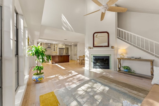 unfurnished living room featuring a fireplace, light wood-style flooring, a towering ceiling, ceiling fan, and stairs