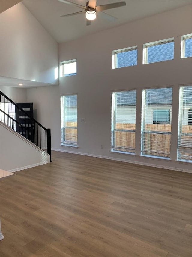 unfurnished living room featuring ceiling fan, a high ceiling, wood finished floors, baseboards, and stairs