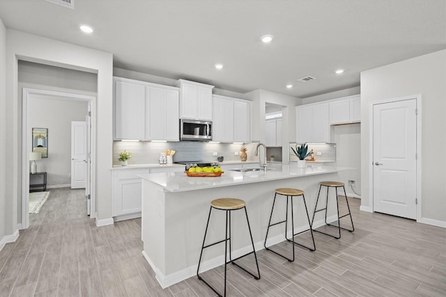 kitchen with a sink, white cabinetry, light wood-style floors, stainless steel microwave, and an island with sink