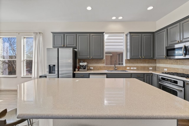 kitchen featuring gray cabinetry, a sink, appliances with stainless steel finishes, backsplash, and a center island