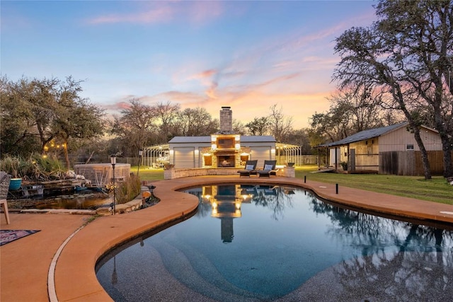 pool at dusk with a warm lit fireplace, an outdoor structure, fence, a yard, and a patio area