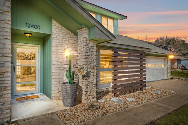 exterior entry at dusk with a shingled roof, stone siding, driveway, and an attached garage