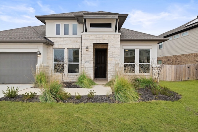 prairie-style home featuring stone siding, roof with shingles, an attached garage, fence, and a front lawn