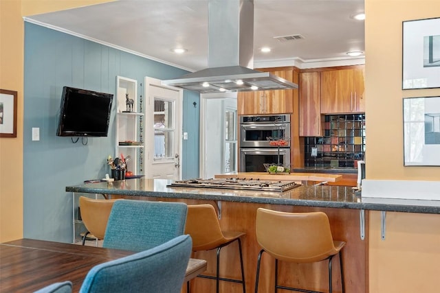 kitchen featuring stainless steel appliances, island exhaust hood, brown cabinets, crown molding, and a kitchen bar