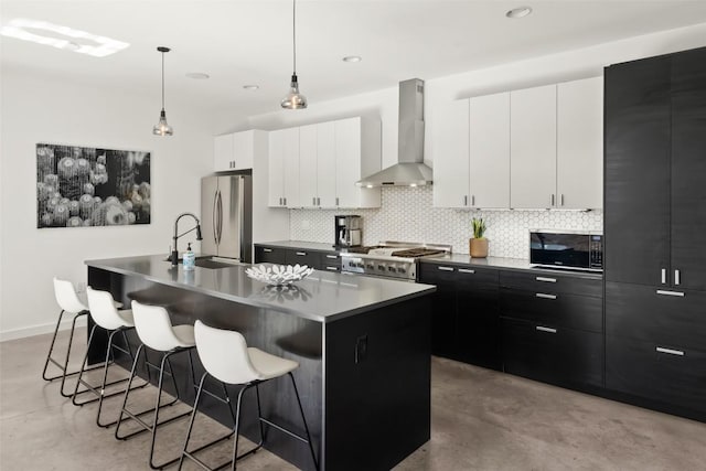 kitchen featuring dark cabinets, stove, wall chimney range hood, backsplash, and freestanding refrigerator