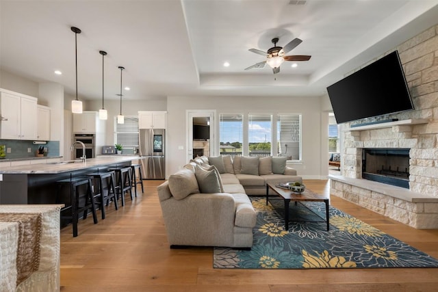 living area featuring a ceiling fan, a tray ceiling, a fireplace, and light wood finished floors