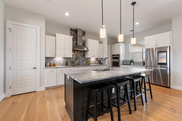 kitchen with a sink, light wood-style floors, appliances with stainless steel finishes, backsplash, and wall chimney exhaust hood