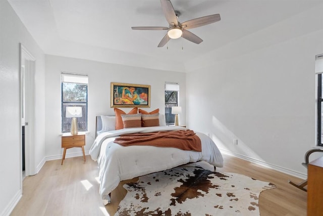 bedroom featuring light wood-type flooring, a raised ceiling, a ceiling fan, and baseboards