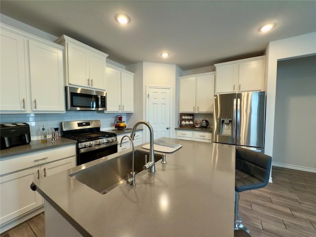 kitchen with wood finished floors, a center island with sink, a sink, stainless steel appliances, and backsplash