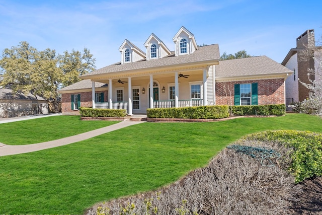 new england style home featuring brick siding, roof with shingles, a porch, a front yard, and ceiling fan