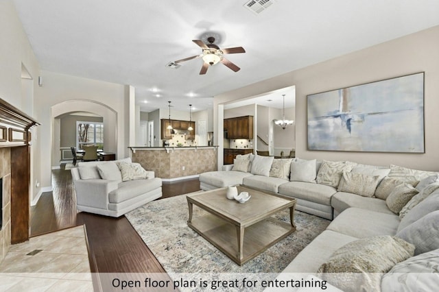 living room featuring arched walkways, light tile patterned floors, visible vents, a tiled fireplace, and ceiling fan with notable chandelier