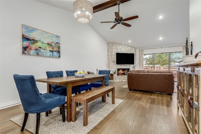 dining room with ceiling fan, high vaulted ceiling, a stone fireplace, wood finished floors, and beam ceiling
