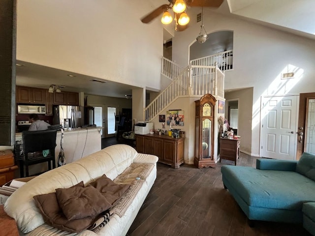 living area featuring dark wood-type flooring, a ceiling fan, a towering ceiling, and stairs