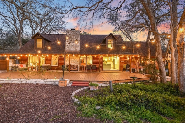back of house at dusk with a deck, roof with shingles, a chimney, and log veneer siding