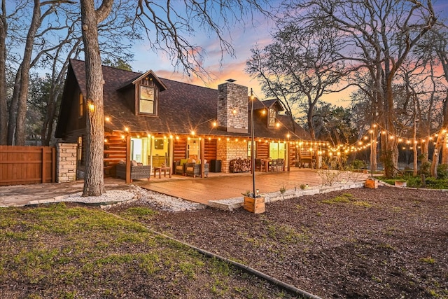back of house at dusk with a shingled roof, fence, stone siding, a chimney, and a patio area