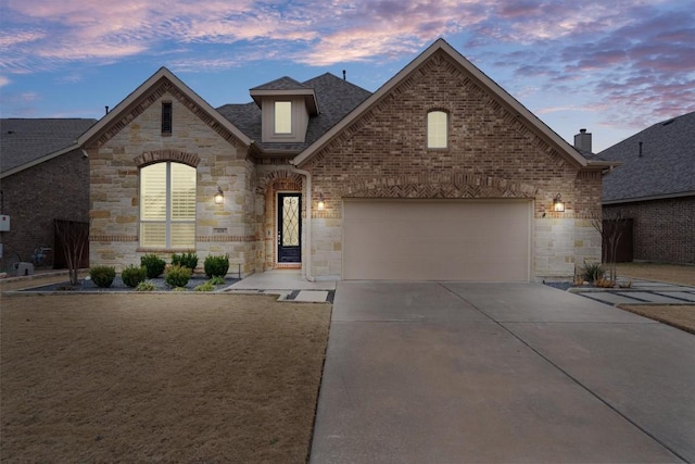 french country inspired facade featuring concrete driveway, stone siding, roof with shingles, an attached garage, and brick siding