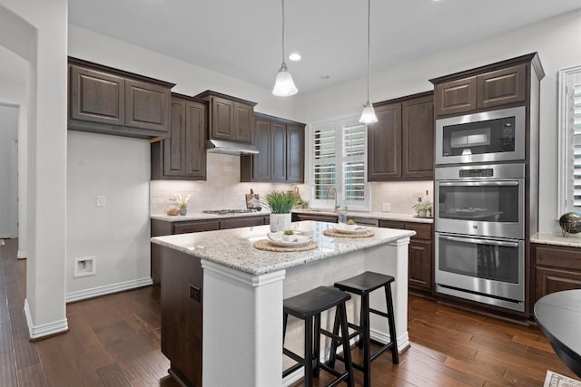 kitchen featuring dark brown cabinetry, under cabinet range hood, stainless steel appliances, decorative backsplash, and dark wood finished floors