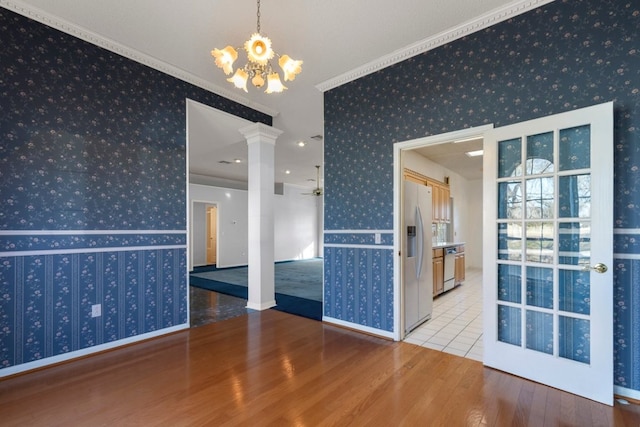 empty room featuring crown molding, wallpapered walls, wood finished floors, ornate columns, and ceiling fan with notable chandelier