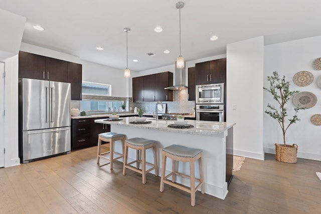 kitchen with a breakfast bar, light wood-style floors, appliances with stainless steel finishes, wall chimney exhaust hood, and tasteful backsplash