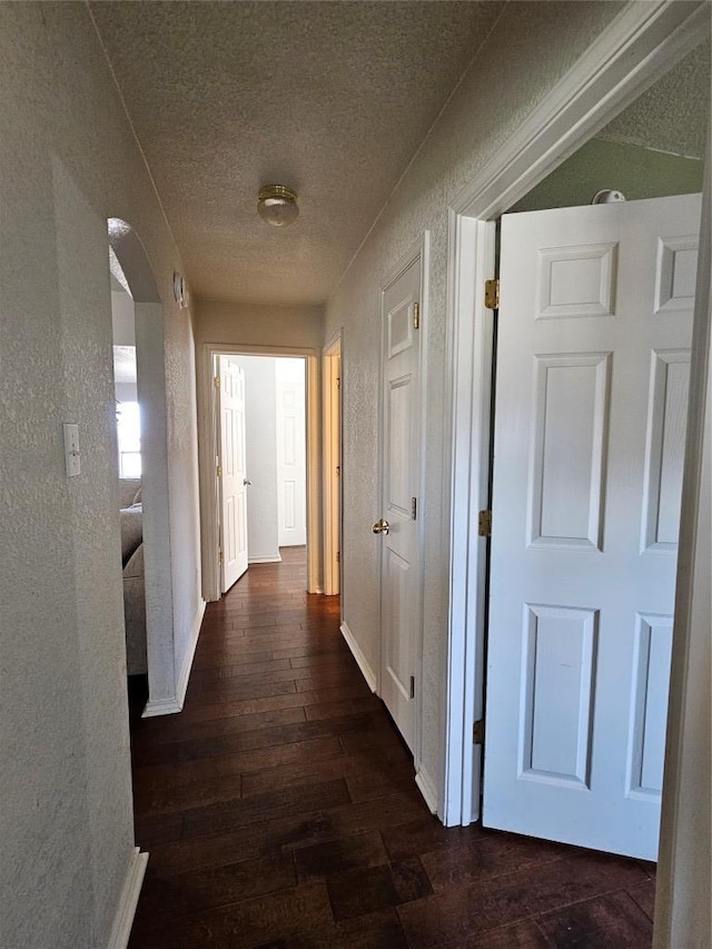 corridor with arched walkways, dark wood-style flooring, a textured ceiling, and baseboards