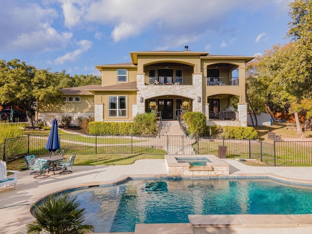 rear view of property with a balcony, stone siding, an in ground hot tub, fence, and stucco siding