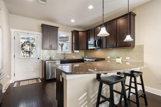 kitchen featuring dark wood-style flooring, appliances with stainless steel finishes, dark brown cabinetry, dark stone counters, and a peninsula