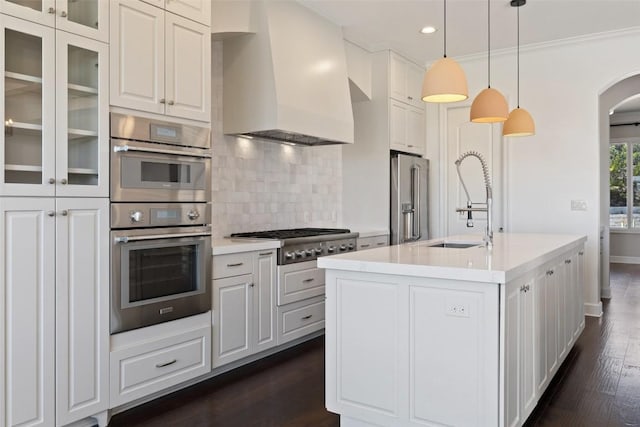 kitchen with arched walkways, wall chimney exhaust hood, appliances with stainless steel finishes, dark wood-style flooring, and a sink