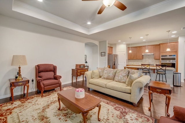 living room with baseboards, arched walkways, ornamental molding, a tray ceiling, and light wood-type flooring