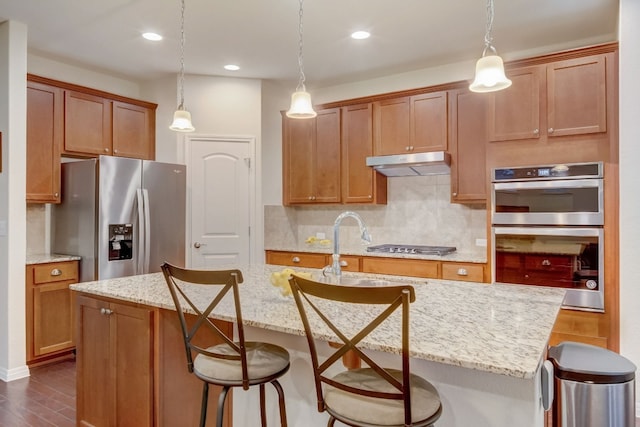 kitchen featuring dark wood-style flooring, decorative backsplash, appliances with stainless steel finishes, an island with sink, and under cabinet range hood