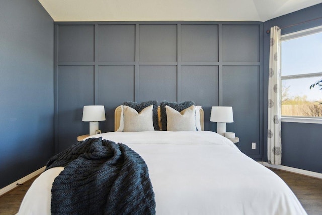 bedroom with dark wood-style floors, vaulted ceiling, baseboards, and a decorative wall