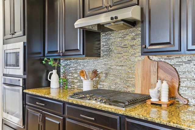 kitchen with light stone counters, under cabinet range hood, dark brown cabinets, appliances with stainless steel finishes, and backsplash