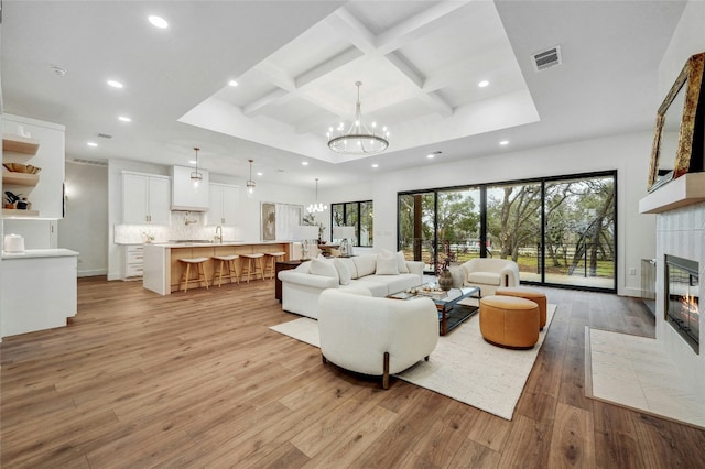 living area featuring a fireplace, coffered ceiling, visible vents, light wood-style floors, and an inviting chandelier